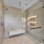 This image depicts a modern, minimalist shower space with a clean and elegant design. The shower area features large beige tiles covering the walls and floor, creating a cohesive and sophisticated look.
1. **Shower Fixtures:** The showerhead is a sleek, modern rain shower type, mounted high on the wall. Adjacent to it is a hand-held shower wand with a simple, streamlined design.
2. **Bench:** A floating bench made of light-colored stone or composite material is installed along one wall, provi