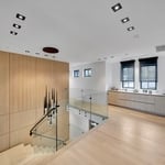 The space showcases a sophisticated modern design with light wooden flooring that extends throughout the area, creating a warm and inviting ambiance. A prominent feature is the glass railing that lines the staircase, which adds to the open and airy feel of the space. The staircase itself is composed of light wood steps, seamlessly blending with the flooring. On the wall adjacent to the stairs, there is a large wooden panel, contributing to the clean and cohesive aesthetic. The ceiling is fitted