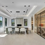 This image depicts a modern and well-lit dining area with a sleek and stylish design. The room exudes a contemporary elegance, marked by its clean lines and sophisticated features.
1. **Dining Table:** The central feature is a large rectangular dining table with a solid surface, surrounded by eight chairs with white upholstery, providing ample seating for gatherings.
2. **Lighting:** Above the dining table hangs a modern chandelier with an industrial design, featuring multiple light bulbs susp