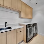 This image depicts a modern, well-organized laundry room with a clean and functional design. The room is designed to maximize efficiency and storage while maintaining a sleek and minimalist aesthetic.
1. **Cabinetry:** Light wood cabinets are installed above and below the countertop, providing ample storage space for laundry supplies and other essentials. The upper cabinets extend across the wall, offering plenty of room to keep items neatly tucked away.
2. **Countertop:** The countertop is ma