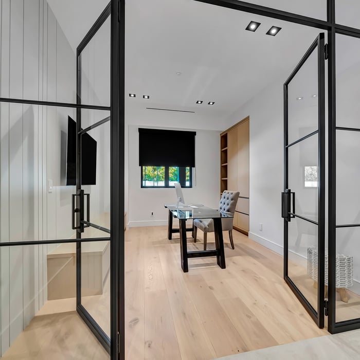 This image shows a modern office space characterized by its minimalist design and functional layout.
1. **Glass Doors:** The office is enclosed with black-framed glass doors, providing a stylish and transparent barrier that maintains privacy while allowing light to flow through.
2. **Desk and Chair:** Inside the office, there is a sleek glass desk paired with a cushioned office chair, creating a functional and stylish workspace.
3. **Wall-Mounted TV:** A flat-screen TV is mounted on one wall,