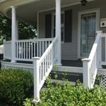 Front view of the covered wrap around porch, with white porch railings and columns. The lattice work below the porch helps to give a backdrop for the shrubbery.
