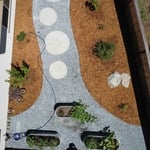 Gravel pathway with circular concrete steps with metal edging. Three planters for vegetables with drip system. Large decorative rocks installed. Planted fruit and decorative plants. Shredded bark for mulch.